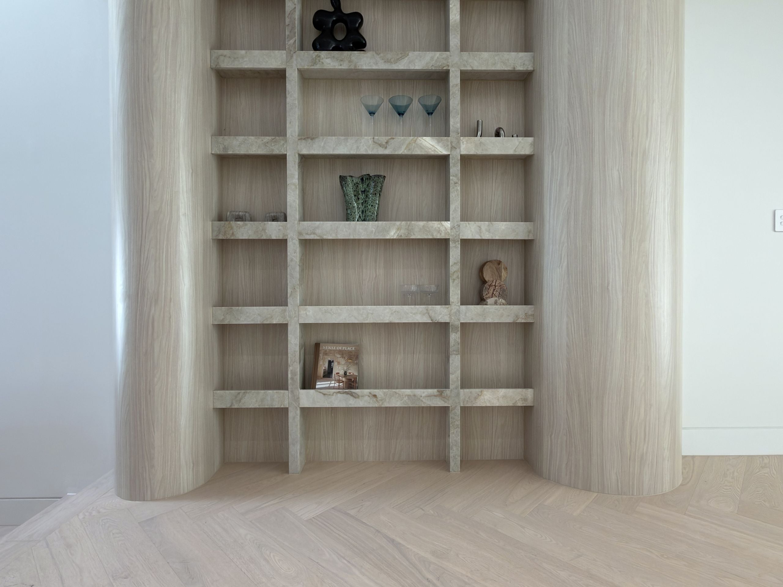 A light wood and marble built-in shelving unit with a grid of open cubbies, styled with small decorative objects, set in a minimalist room with light wood flooring.