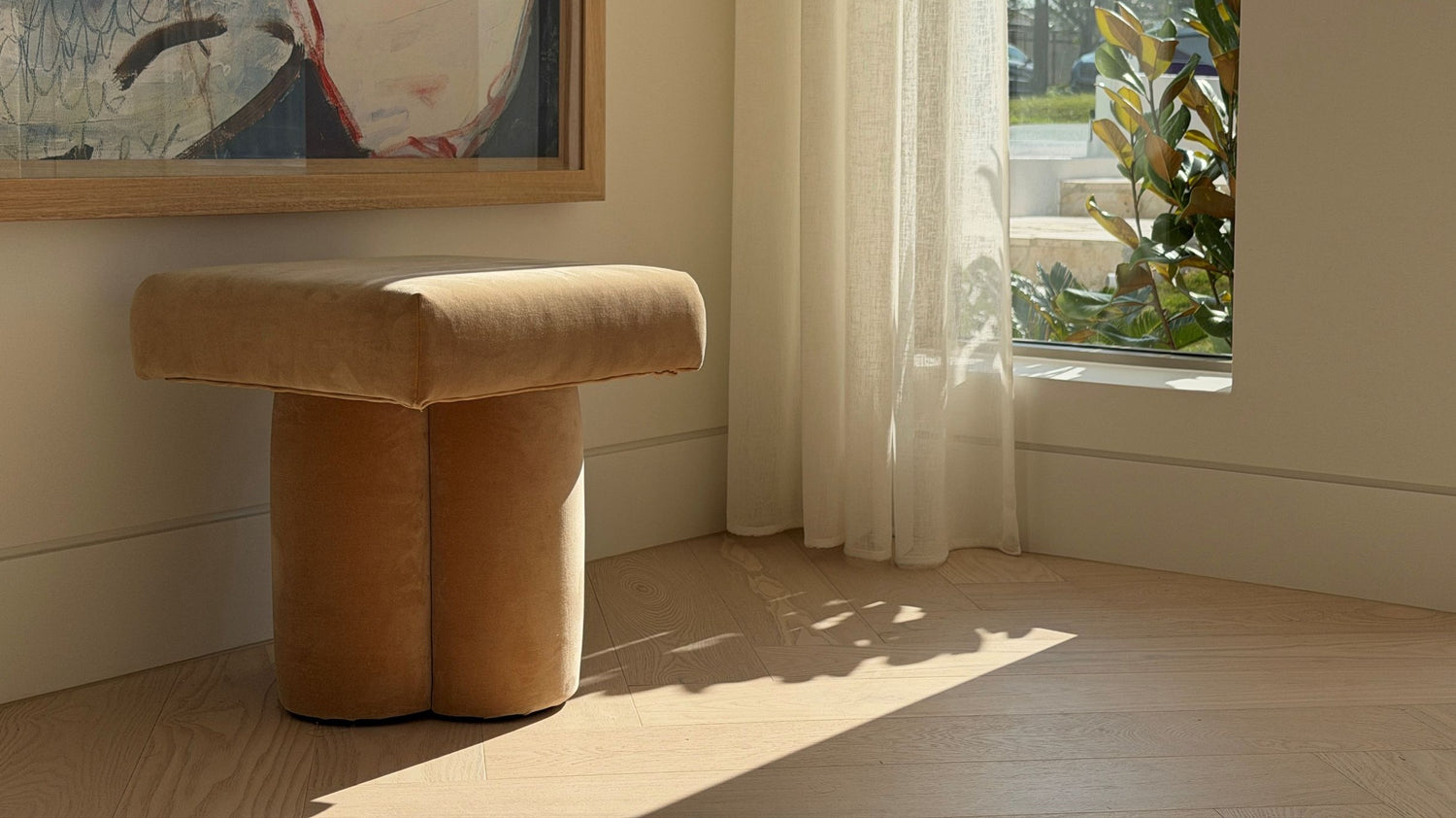 A modern upholstered stool with a thick rectangular seat and two cylindrical legs in a warm tan velvet fabric, placed in a sunlit room with a window, sheer curtains, and a framed painting on the wall.