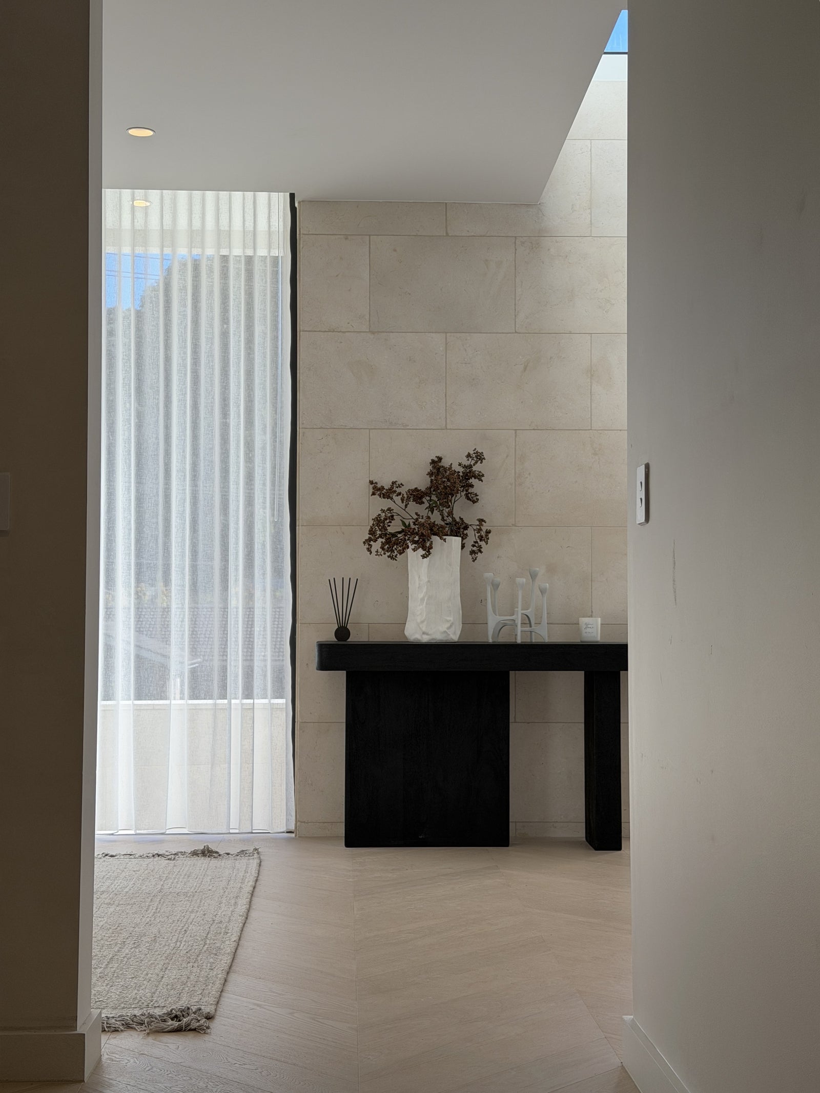 A modern interior hallway with a black console table against a light beige tiled wall, holding a white vase with dried branches and glassware. A sheer white curtain hangs by a window on the left, and a light wood floor and rug are visible.