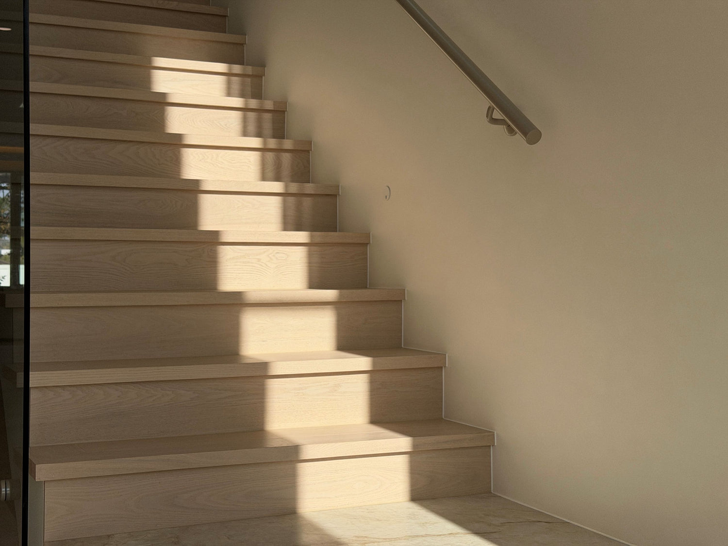 Light natural wood staircase steps with a dark metal handrail against a warm beige wall, lit by directional sunlight that casts strong shadows.