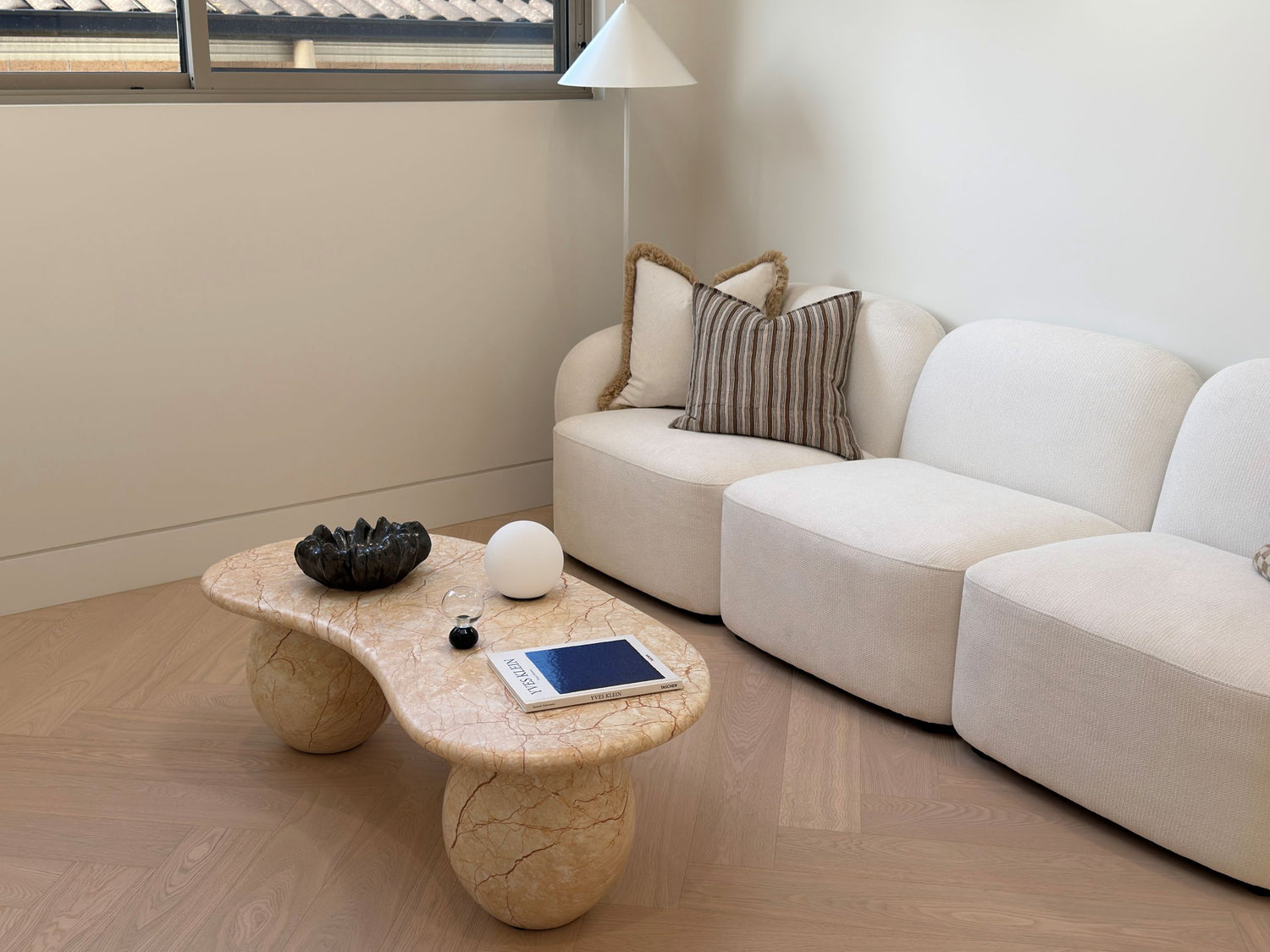 A modern living room corner featuring a curved beige stone coffee table with chunky legs, styled with a black lotus bowl, white orb, and tablet, next to a cream-colored modular sofa with decorative pillows.
