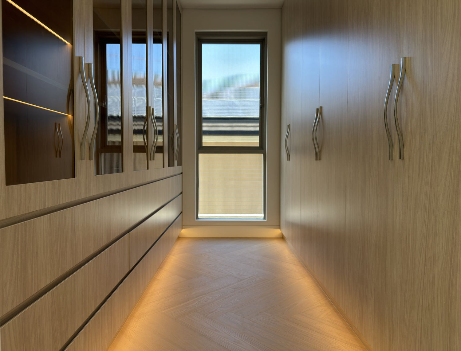 A modern walk-in closet with light wood cabinets, silver wavy handles, and a herringbone wood floor, featuring a central window looking out onto a balcony.