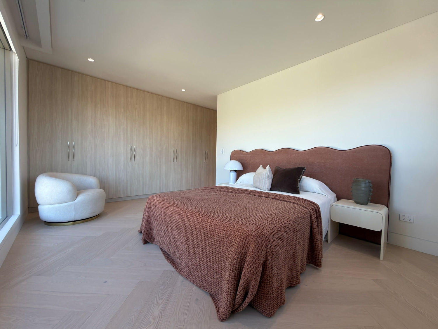 A modern bedroom featuring a bed with a textured rust-brown blanket and matching headboard, set against cream walls with a built-in wood wardrobe and a white accent chair.