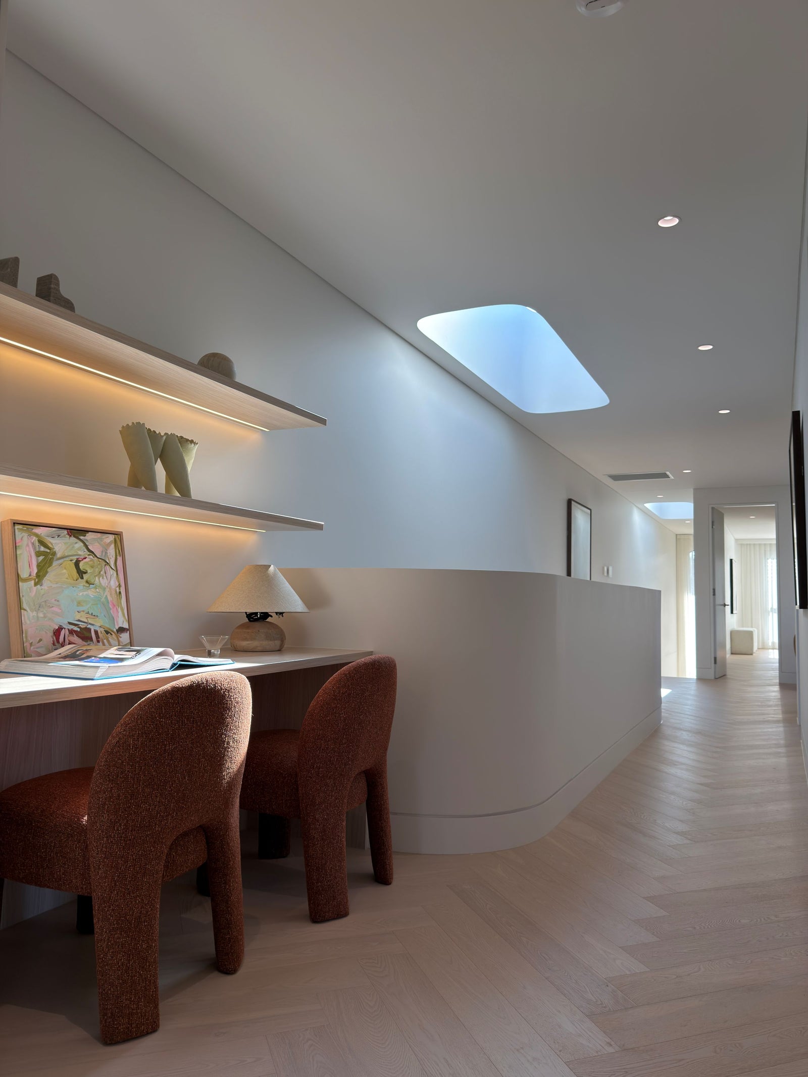 A modern interior hallway with light wood flooring, white walls, built-in floating shelves, a small desk, a table lamp, and two textured rust-brown upholstered chairs.
